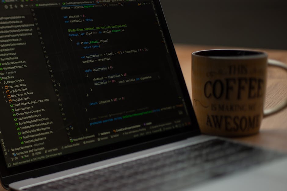 A person operating a laptop on a dark surface, with visible code or programming language on the screen, possibly in a home or office setting. The individual is using both hands, one on the keyboard an